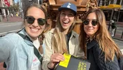 Three happy individuals are posing for a selfie on a sunny day, with one of them holding a book that has a yellow post-it note on its cover.