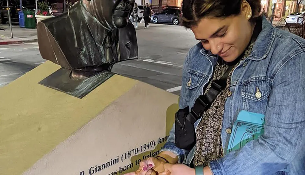 A person is smiling while seemingly shaking hands with a bronze statues hand which is extending out from a wall plaque