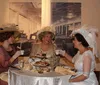 Three women dressed in vintage clothing with elaborate hats are having a cheerful conversation in an elegantly furnished room