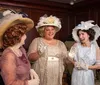 Three women dressed in vintage clothing with elaborate hats are having a cheerful conversation in an elegantly furnished room