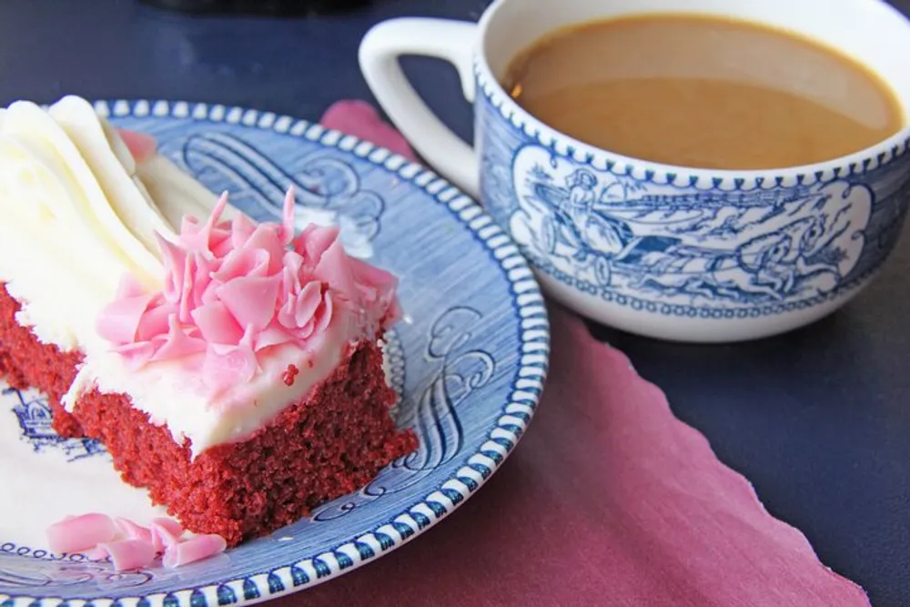 A piece of red velvet cake topped with cream cheese frosting and pink sprinkles is served on a decorative blue and white plate beside a matching cup filled with coffee