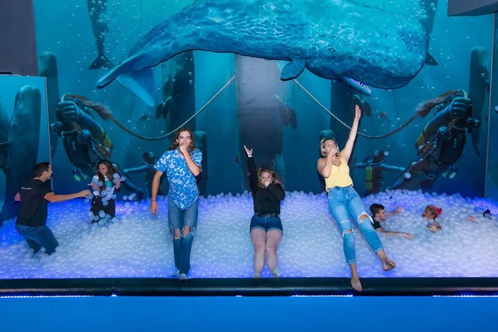 Visitors are enjoying an immersive experience in a ball pit with a simulated underwater setting complete with a large whale model and sea creature exhibits in the background