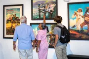 Three individuals are attentively viewing a collection of vibrant paintings at an art gallery.