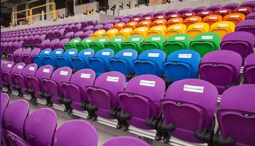 The image shows a colorful array of empty seats in a stadium or auditorium with each seat displaying a sign indicating social distancing measures