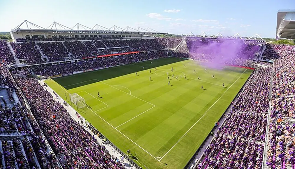 The image shows a vibrant soccer stadium filled with spectators with one end engulfed in a large cloud of purple smoke likely from a smoke bomb or flare set off by fans