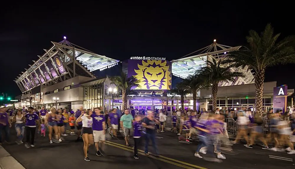 Crowds of people are gathered outside a stadium adorned with a large lion emblem in the evening