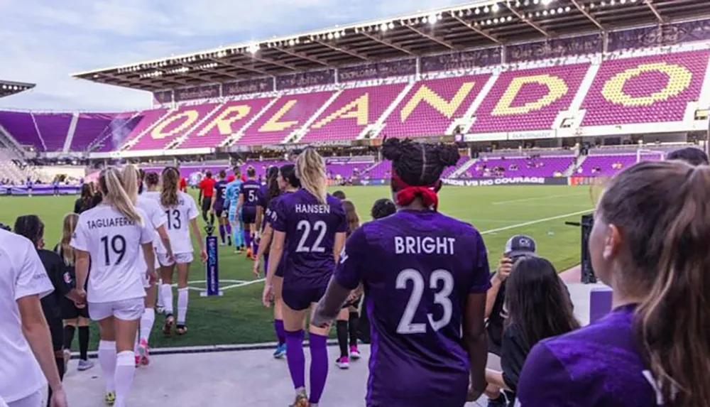 Players are walking onto a soccer pitch in a stadium with ORLANDO displayed across the seating