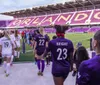 Soccer players are walking onto a stadium with purple seating and the word ORLANDO prominently displayed in the background