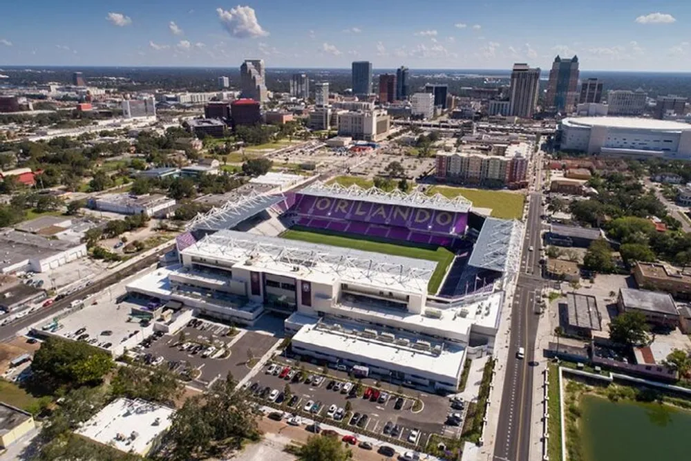 This image shows an aerial view of a stadium with ORLANDO written on the seats surrounded by urban development and roadways under a partly cloudy sky