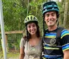 Two individuals are mountain biking on a forest trail surrounded by lush greenery