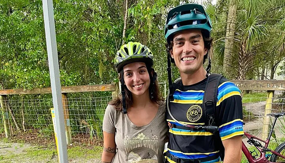 Two happy cyclists wearing helmets are posing for a photo with a bike partially visible in the background