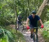 Two individuals are mountain biking on a forest trail surrounded by lush greenery