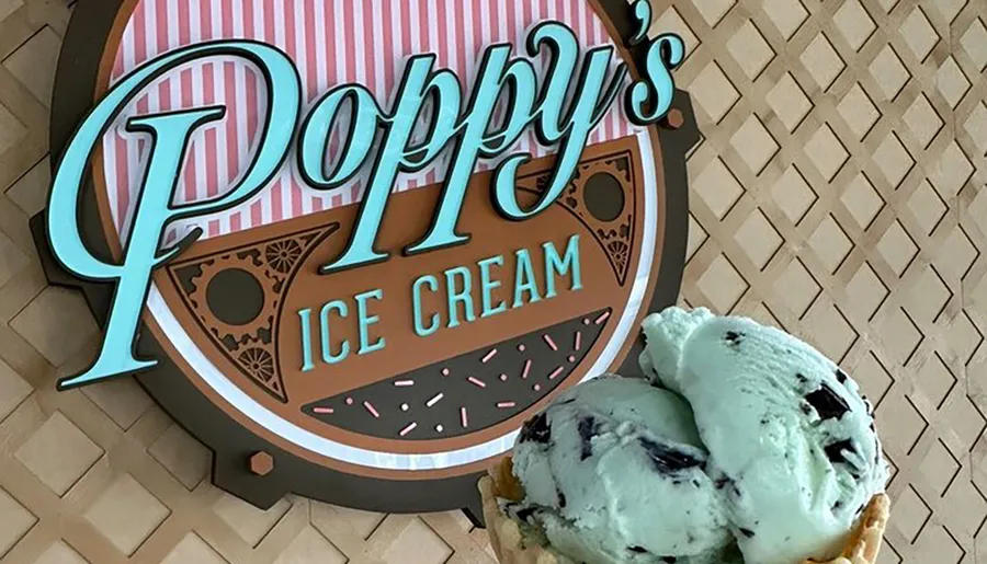 A scoop of mint chocolate chip ice cream in a cone is showcased against the background of a Poppy's Ice Cream shop sign.