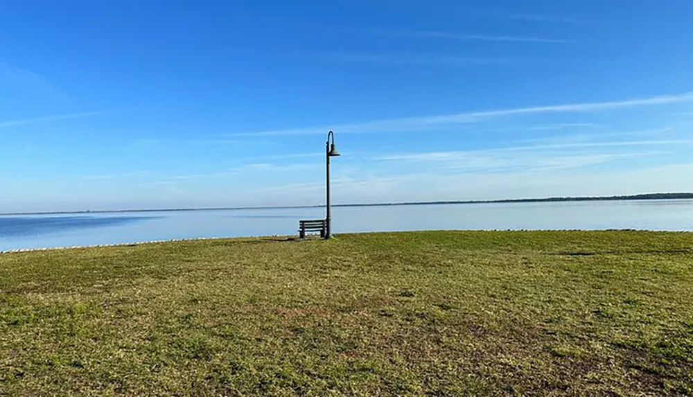 A solitary bench with a lamp post nearby sits facing a vast expanse of calm water under a clear sky