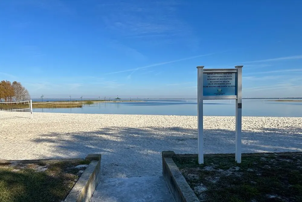 The image shows a tranquil lakeside scene with a sandy shore a volleyball net and a sign that likely contains information about the area