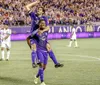 Two soccer players are celebrating during a match with one player riding piggyback on the other against the backdrop of a stadium filled with fans