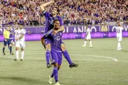 Two soccer players are celebrating during a match, with one player riding piggyback on the other against the backdrop of a stadium filled with fans.