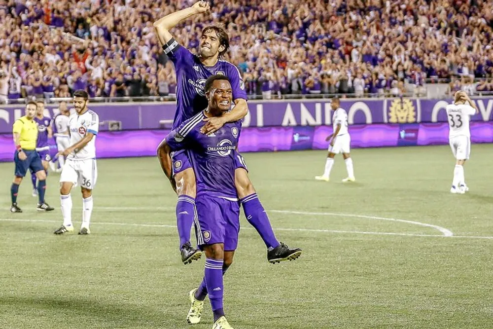 Two soccer players are celebrating during a match with one player riding piggyback on the other against the backdrop of a stadium filled with fans