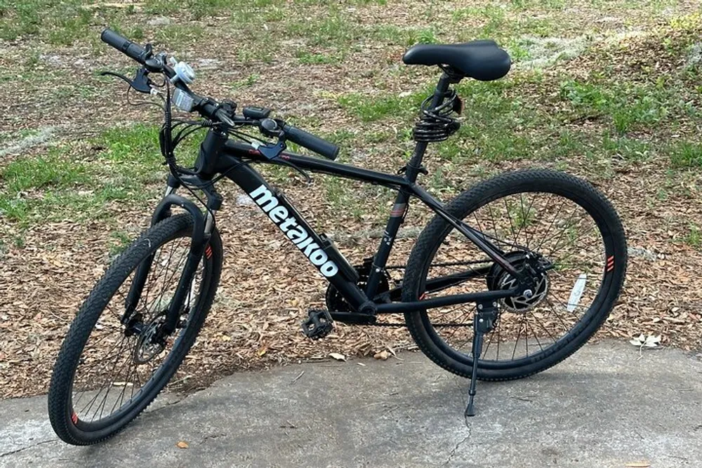 A black mountain bike is parked outdoors on a concrete slab surrounded by grass and leaf litter