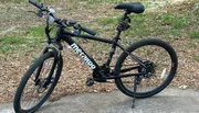 The image shows a black mountain bike with the brand name 'Metakoo' displayed on its frame, stationed outdoors on a concrete surface with grass and fallen leaves in the background.