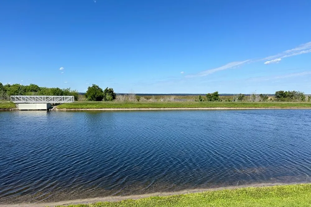 The image shows a serene body of water with a metal pedestrian bridge to the left grassy banks and a clear blue sky