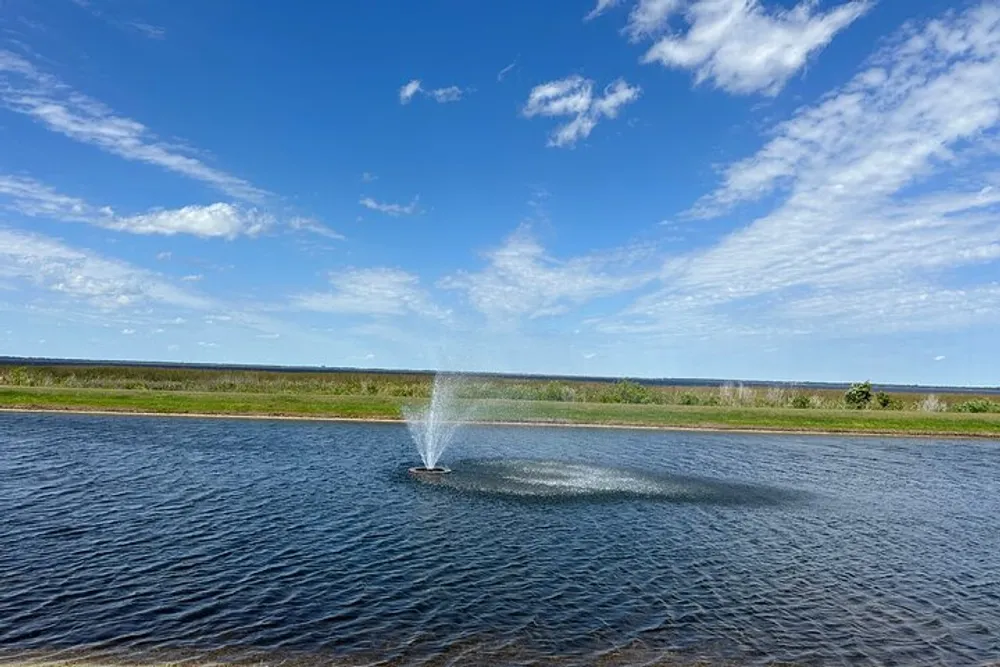 A water fountain sprays into the air in the middle of a placid lake under a blue sky dotted with white clouds