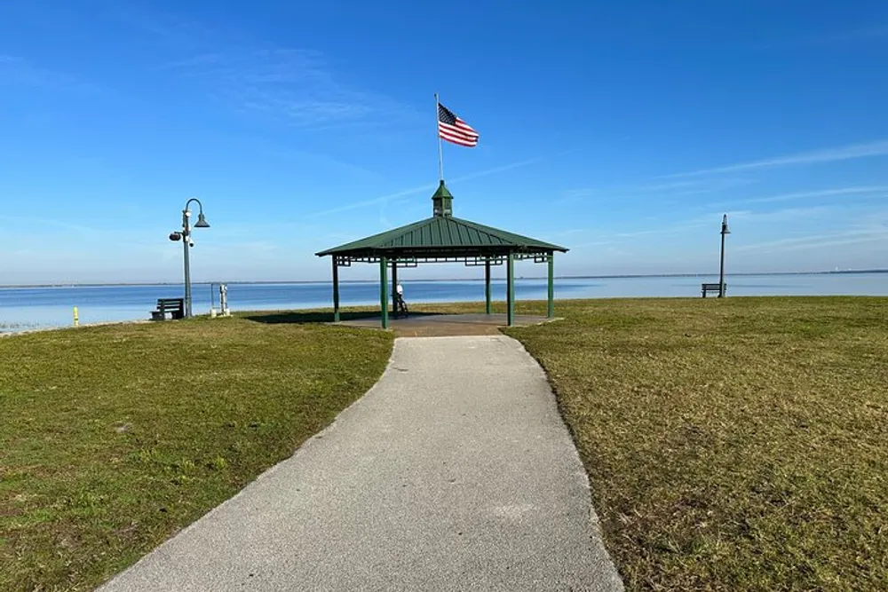 A paved pathway leads to a gazebo flying an American flag overlooking a calm body of water under a clear blue sky