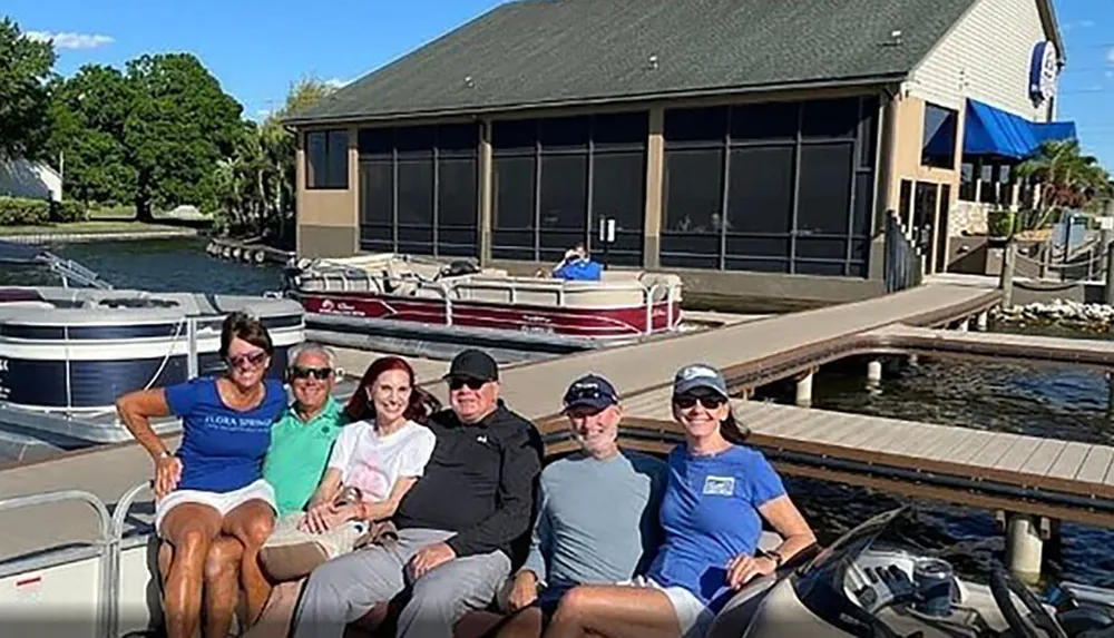 A group of six people is smiling for the camera while seated on a docked pontoon boat in front of a building on a sunny day