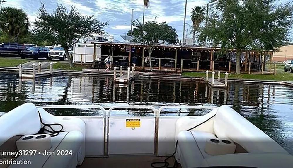 The image shows the back of a white pontoon boat on the water with a view of a dock and a waterfront dining area adorned with string lights surrounded by trees and vehicles parked nearby