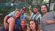 A group of five cheerful adults poses for a lighthearted photo outdoors, with a bridge and foliage in the background.