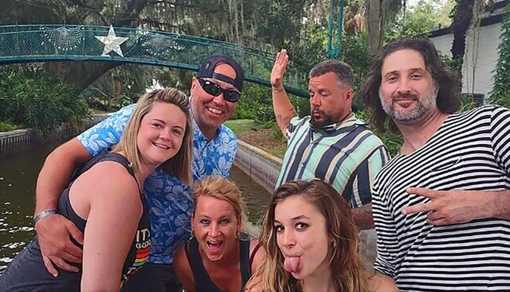 A group of five cheerful adults poses for a lighthearted photo outdoors with a bridge and foliage in the background