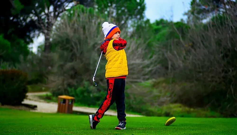 A child in colorful winter clothes is holding a golf club mid-swing on a lush green course