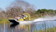 A group of people are enjoying a fast ride on an airboat cutting across what appears to be a calm river or wetland area.