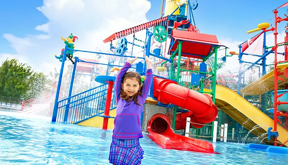 A cheerful child is raising her arms in delight at a colorful water playground with slides and water features