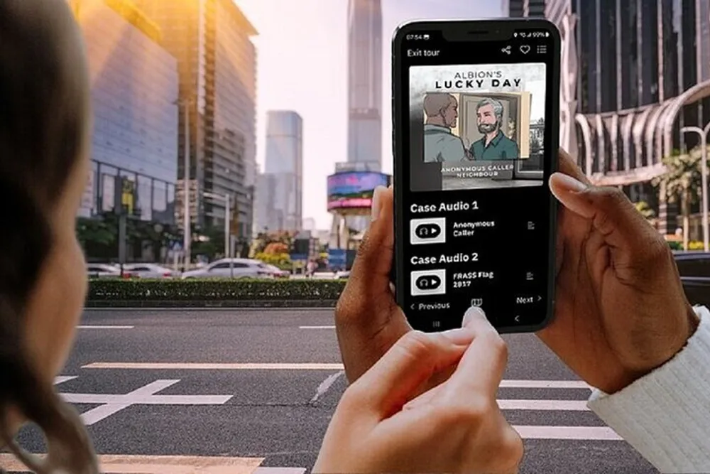 A person is holding a smartphone displaying a screen with an audio tour app called Albions Lucky Day with multiple episodes available while standing at a pedestrian crossing in a city