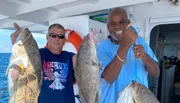Two smiling men are posing on a boat, each holding up a large fish they seemingly caught.