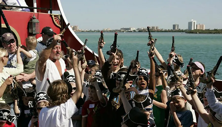 A group of excited children and adults, some wearing pirate hats, raise their toy guns in the air on a pirate-themed boat ride.