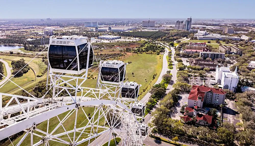 An aerial view of a large observation wheel with enclosed cabins overlooks a scenic area with trees roads buildings and water bodies in the distance