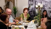 A group of people enjoys a lively dinner party, with one woman in the center laughing heartily.