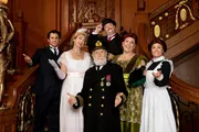 A group of people dressed in early 20th century attire, representing characters from the Titanic era, pose cheerfully on an ornate wooden staircase.