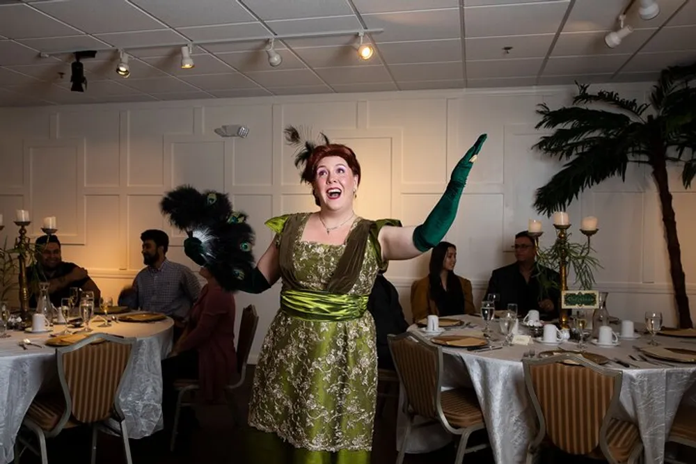 A performer in a green vintage dress and feathered hairpiece is enthusiastically gesturing with a fan in her hand at a dining event