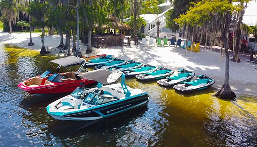 The image shows a variety of watercraft, including boats and jet skis, moored near a sandy beach area with trees and some colorful beach chairs, suggesting a recreational water activity spot.