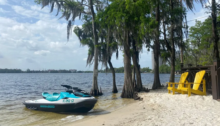 A personal watercraft is moored on the sandy shore of a lake with Spanish moss-draped trees, next to a pair of inviting yellow Adirondack chairs.