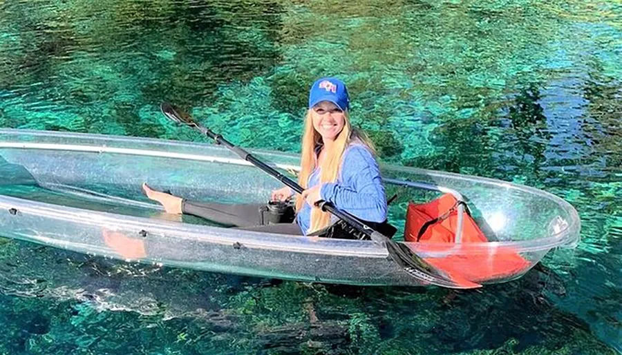 A person is smiling while sitting in a transparent kayak over clear, turquoise water.