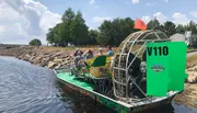 An airboat with several passengers is ready for a tour on a calm waterway, under a partly cloudy sky.