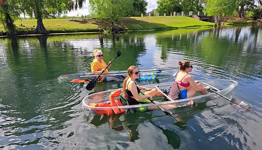 Three people are paddling in transparent kayaks on a calm body of water, offering a clear view of the underwater scene.