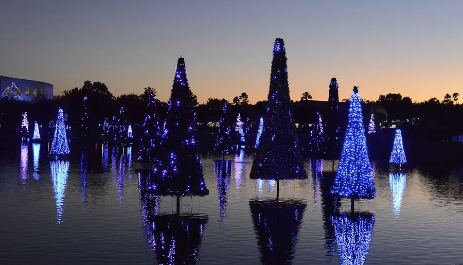 The image shows a serene twilight scene with multiple blue-lit Christmas trees reflected in a body of water, creating a tranquil and festive atmosphere.