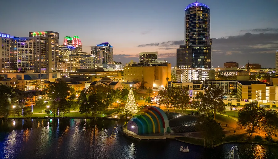 The image captures a twilight cityscape featuring a lit Christmas tree near a colorful, dome-shaped structure with a waterfront park, and a backdrop of illuminated high-rise buildings.