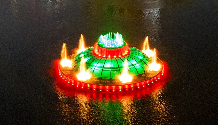 The image shows an aerial view of a brightly lit fountain with a green dome center and surrounding fiery spouts, possibly indicating a nighttime light and water show.