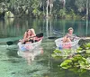 Four people are smiling and posing with their paddles in clear kayaks on a calm transparent waterway amidst lush greenery